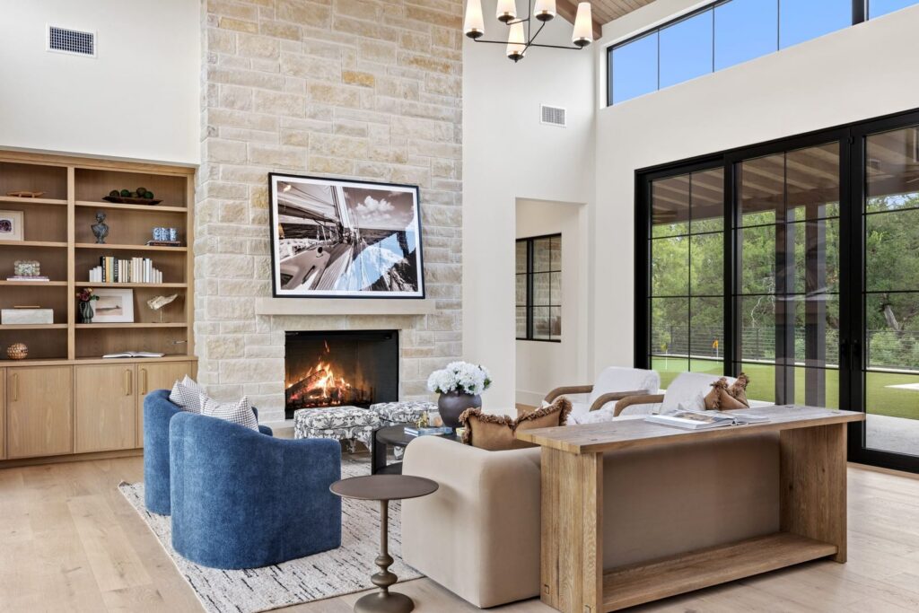 Bright modern living room with a stone fireplace, framed photo above the mantel, built-in wooden shelves, blue armchairs, beige sofa, and a wooden console desk near large black-framed sliding doors opening to a green yard