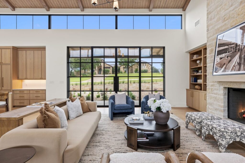 Open-concept living room with a beige sofa, two blue armchairs, and a round coffee table in front of large black-framed glass doors opening to a green yard.