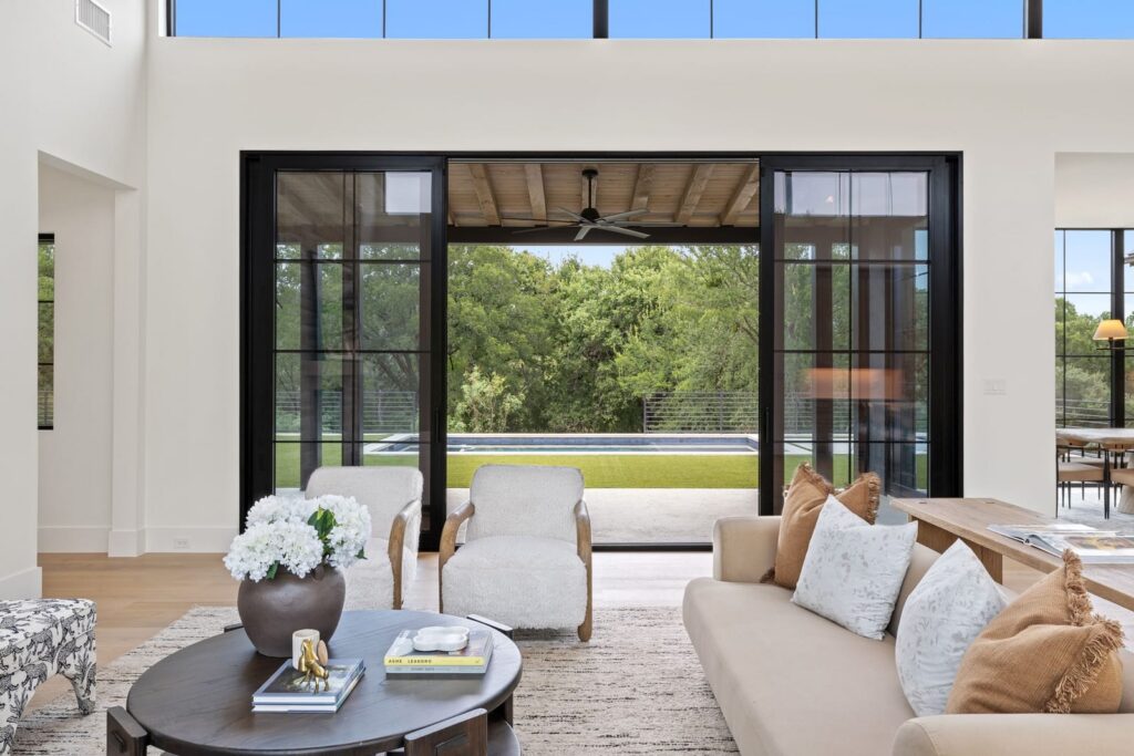 Bright living room with a round coffee table, white hydrangeas, and a beige sofa facing large black-framed glass doors opening to a pool yard outside.
