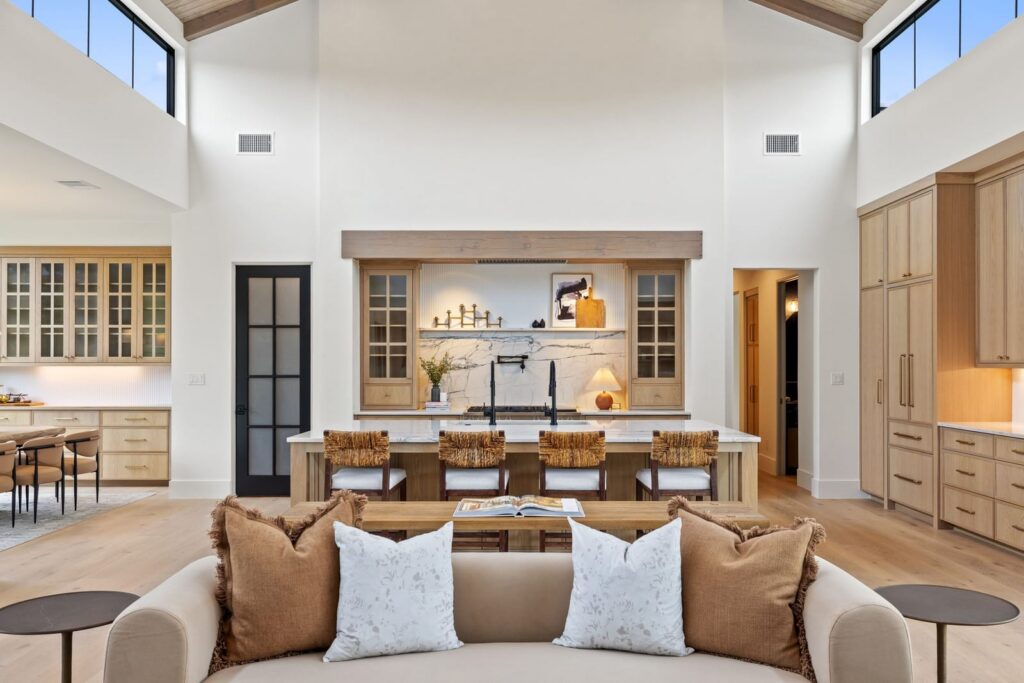 Open-concept kitchen and living area with a marble-topped island, wooden stools, light wood cabinets, and a high vaulted ceiling with exposed beams.