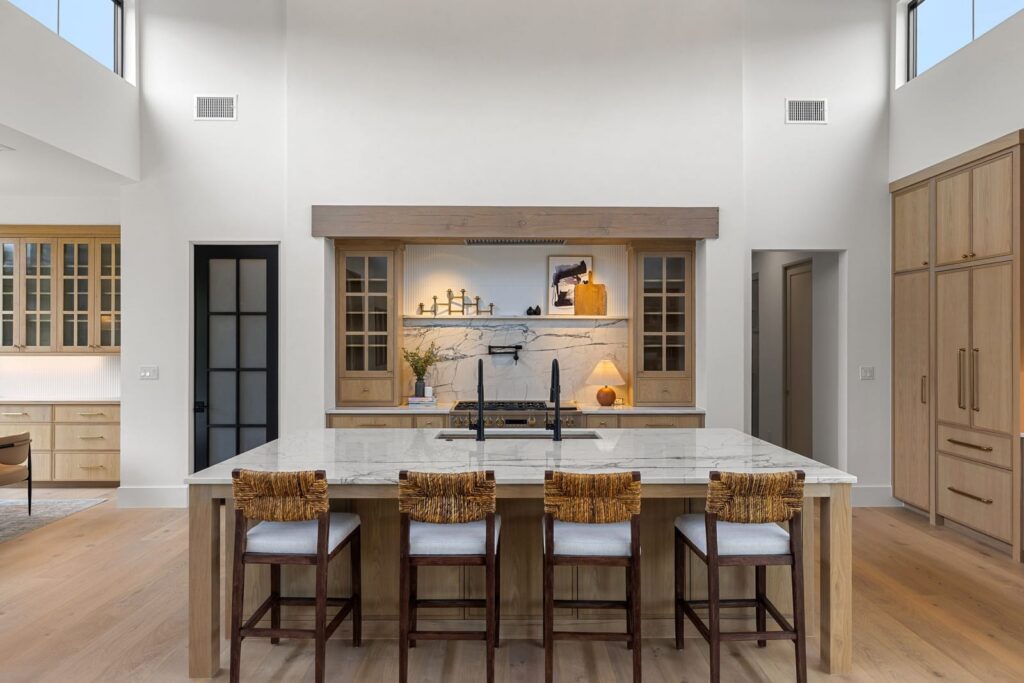 Modern kitchen with a large marble island and four woven-back stools facing a built-in range and marble backsplash.
