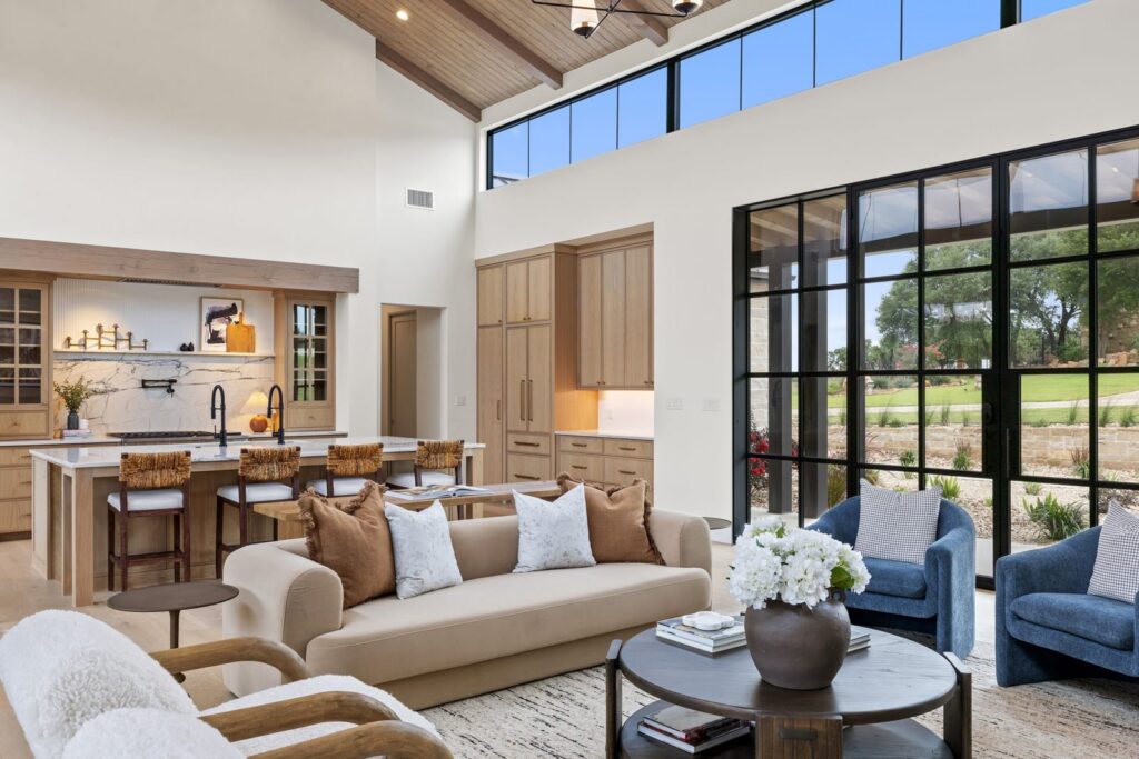Bright open living area with a kitchen island, marble backsplash, and large black-framed windows overlooking a landscaped yard.