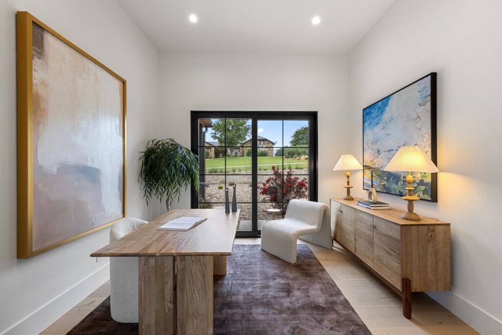 Bright, modern home office with a large wooden table, white chairs, and a sideboard flanked by two lamps, door to a garden outside.
