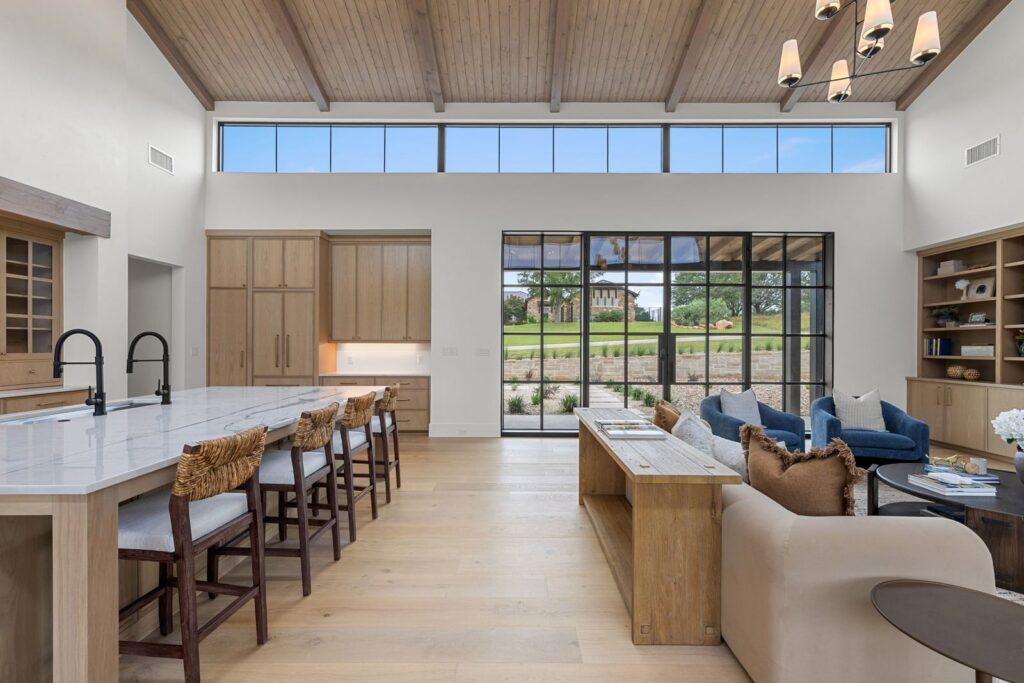 Open-concept kitchen and living room with a marble-topped island, eight bar stools, and light wood cabinetry. A view to the outdoors through large glass doors is visible in the background.