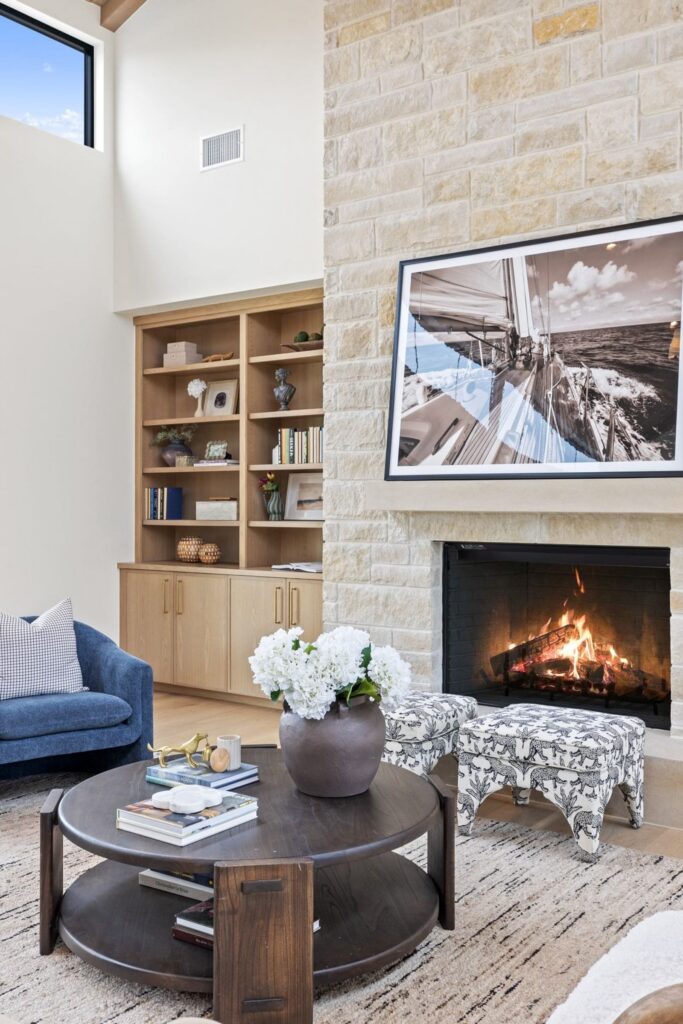 Modern living room featuring a stone fireplace with a large framed sailboat photo, and a wooden bookshelf filled with decor along the left wall.