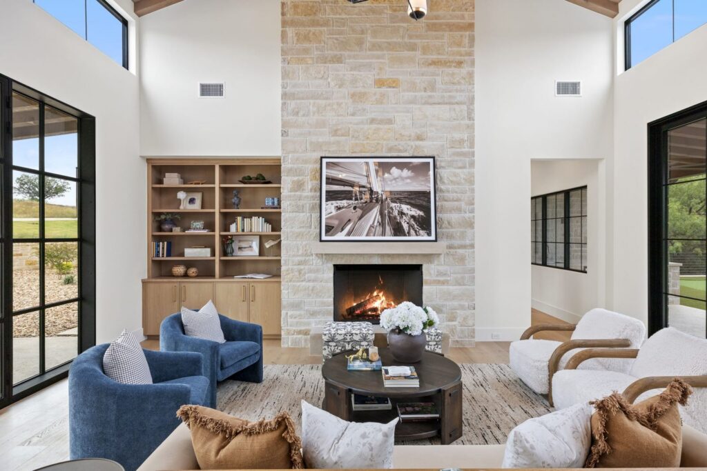 Bright living room with a tall stone fireplace, a framed black-and-white sailboat photo above, built-in wooden shelves, and blue armchairs around a round coffee table.