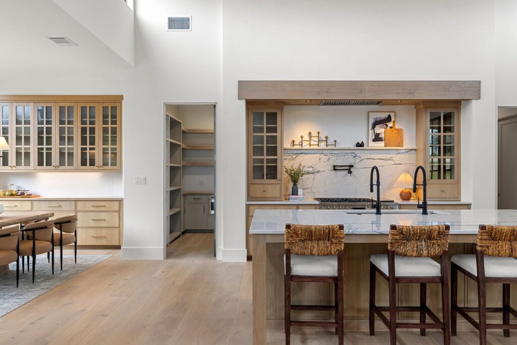 Bright open kitchen with a large marble island, wooden cabinets, and a stainless stove area; built-in pantry to the left and seating at the island in the foreground