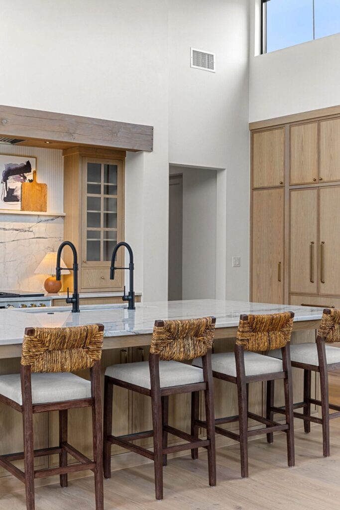 Modern kitchen island with a white marble counter, black dual faucets, and woven-seat bar stools in front of light wood cabinetry.