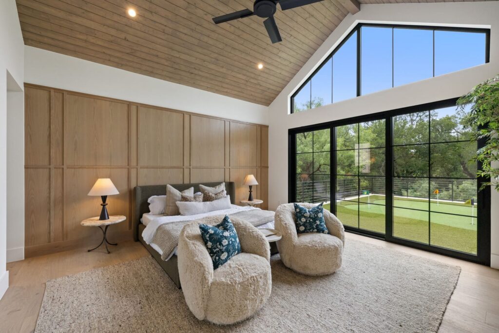 Bedroom with vaulted wood ceiling and large black-framed windows overlooking a green lawn; cozy bed and two fluffy chairs.
