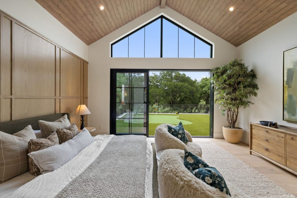 Bright bedroom with a vaulted wooden ceiling, a large bed and plush pillows, and a sliding glass door opening to a green backyard.