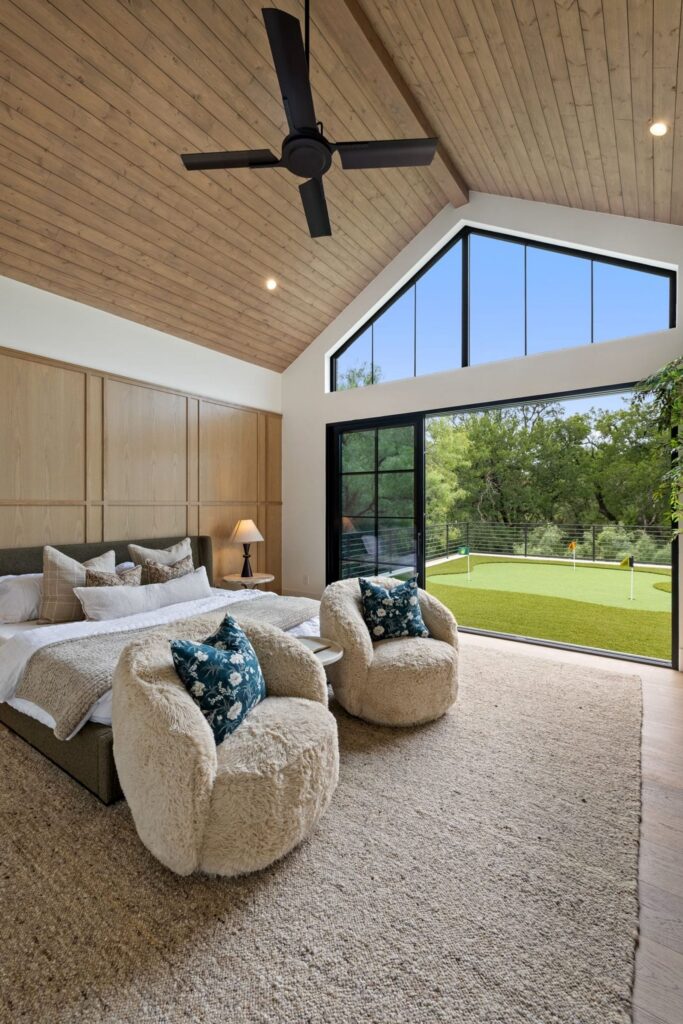 Bedroom with a vaulted wooden ceiling, large black-framed windows, and a sliding glass door opening to a green lawn outside. Plush cream chairs and a bed with neutral bedding and blue patterned pillows sit in the room.