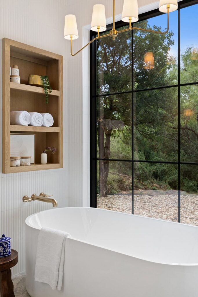 White freestanding bathtub beside a large grid-window, with a gold faucet and a wooden shelf holding towels and decor on a white tiled wall.