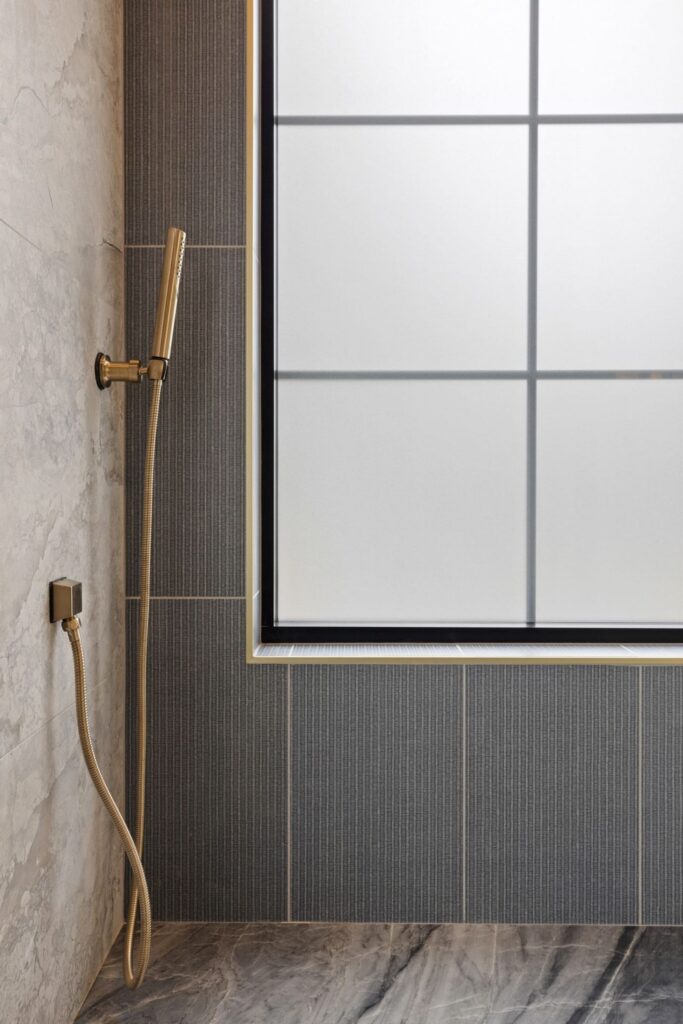 Bronze handheld shower attached to a marble wall beside a frosted grid window on a grey-tiled bathroom wall.