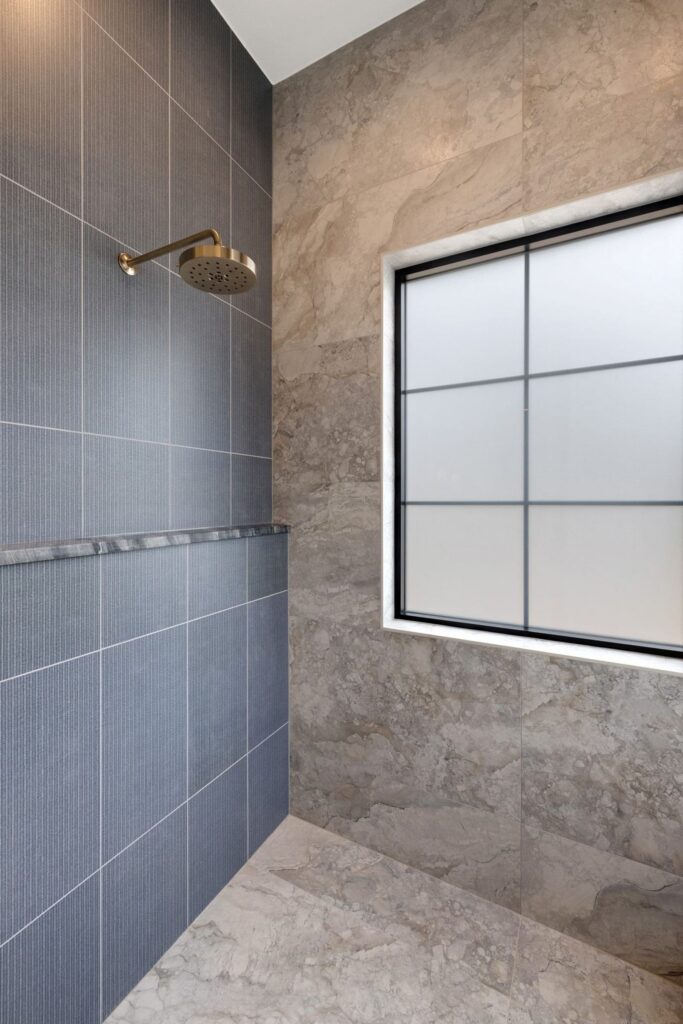 Brass rain showerhead mounted on a dark blue tiled wall in a marble-tiled bathroom beside a frosted grid window.