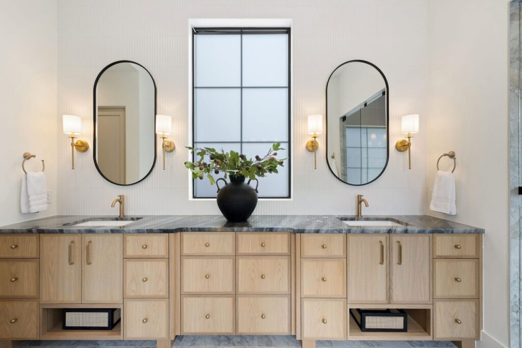 Double vanity with light wood cabinets, gray marble countertops, two black-framed oval mirrors, and gold faucets.