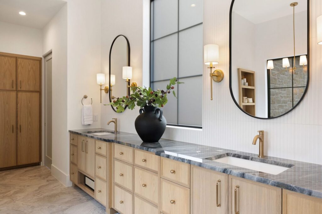 Modern bathroom with light wood cabinetry, marble countertop, and dual sinks beneath round black-framed mirrors. A large black vase with greenery sits on the counter; gold faucets and wall sconces add warmth. A tall wooden storage cabinet is in the background.