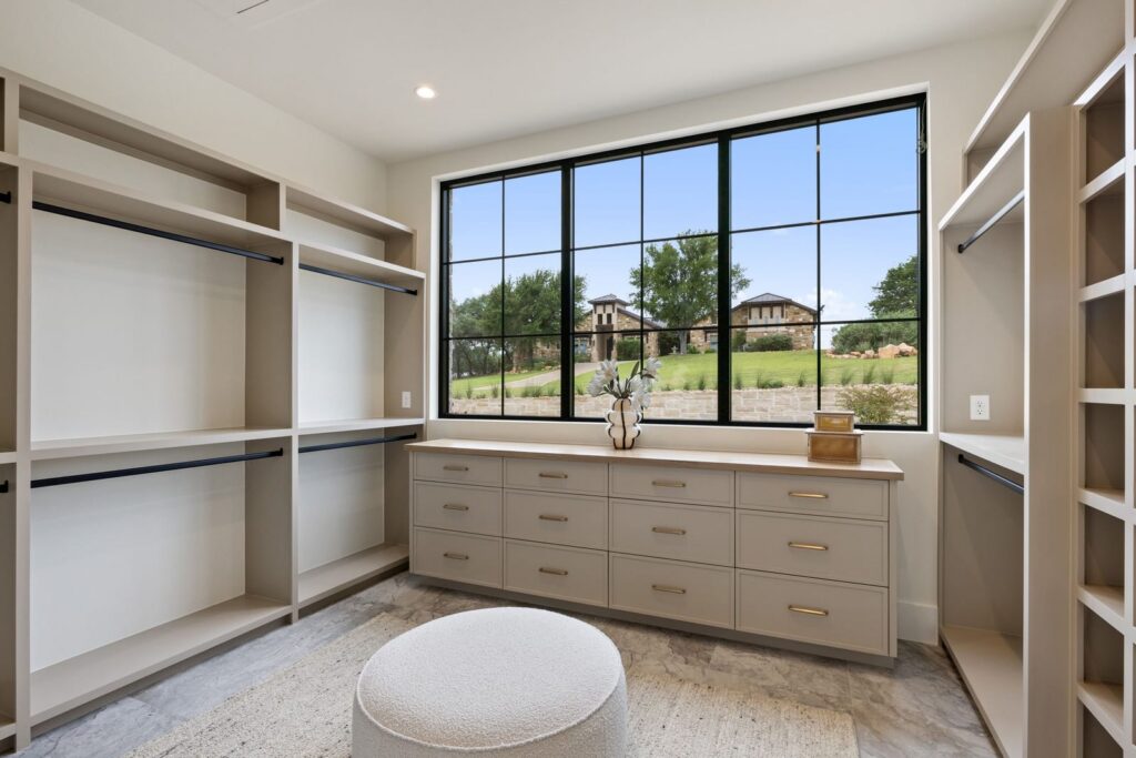 Walk-in closet with open shelves and hanging rods on both sides, a row of light gray drawers under a wide window, and a round ottoman in the center; a vase and small boxes on the countertop create a tidy focal point.